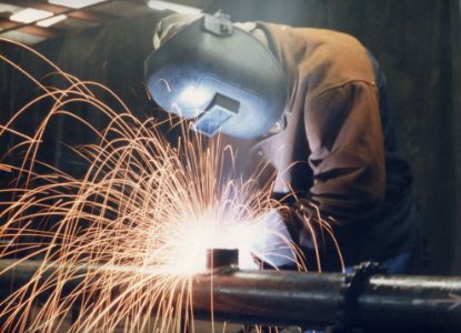 A welder wearing protective gear, including a helmet and jacket, is actively welding a metal pipe. Bright orange sparks fly in all directions as the intense light from the welding process illuminates the scene. The setting appears to be a workshop or industrial environment, with shadows and machinery visible in the background.