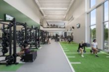 Two-story view inside the Deward and Anne Dopson Fitness Center. The lower level has free weights, strength equipment, and a turf training strip, with people working out. Floor-to-ceiling windows look onto campus, and an adjacent gymnasium with basketball and volleyball courts is visible through interior glass at right.