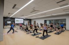 A group fitness class in a studio at the Ferguson Student Union. An instructor at the front demonstrates a lunge while about twenty participants follow along on mats with small exercise balls. A mirrored wall and a projection screen with the Arkansas Tech University logo are behind the instructor.