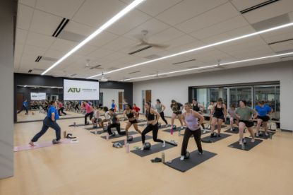 A group fitness class in a studio at the Ferguson Student Union. An instructor at the front demonstrates a lunge while about twenty participants follow along on mats with small exercise balls. A mirrored wall and a projection screen with the Arkansas Tech University logo are behind the instructor.