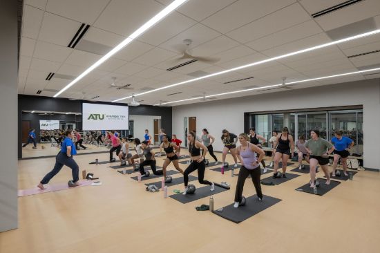 A group fitness class in a studio at the Ferguson Student Union. An instructor at the front demonstrates a lunge while about twenty participants follow along on mats with small exercise balls. A mirrored wall and a projection screen with the Arkansas Tech University logo are behind the instructor.